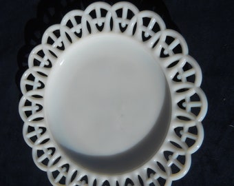 May include: A white, decorative plate with a scalloped, openwork edge. The plate has a smooth, slightly concave center and a detailed, repeating pattern around the rim. The plate is set against a dark background, highlighting its delicate design.