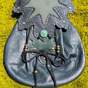 May include: A dark green leather pouch featuring a textured black leather leaf-shaped flap with silver-toned rivets. The pouch is secured with a black cord drawstring, accented by metallic beads and a circular green button with a dragonfly emblem.
