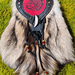 Op de afbeelding: Een handgemaakte bontzak met een rode leren flap met een wolfontwerp. De tas heeft een zwarte leren rand met messing studs, een benhalsketting en zwarte trekkoorden versierd met rode en gouden kralen. De vacht is een mix van bruin, beige en wit.