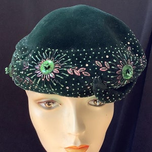 May include: A dark green velvet hat with floral beadwork and sequin accents. The hat features a rounded crown and a decorative bow on the side. The embellishments include green and pink beads and sequins, creating a vintage, elegant look.