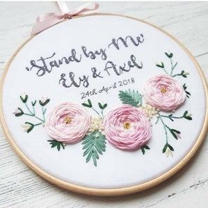 May include: Embroidered hoop art with the text "Stand by Me" and "Ely & Axel" with the date "24th April 2018". The hoop is decorated with pink flowers and green leaves.