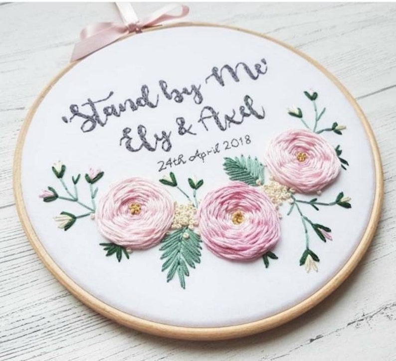 May include: Embroidered hoop art with the text "Stand by Me" and "Ely & Axel" with the date "24th April 2018". The hoop is decorated with pink flowers and green leaves.