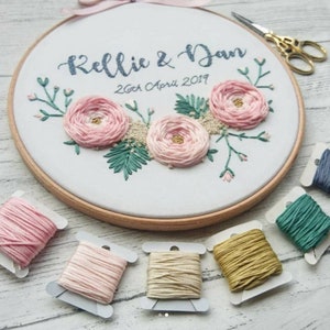 May include: Embroidered hoop art with pink flowers, green leaves, and the text "Kellie & Dan 26th April 2019".  The hoop is surrounded by spools of embroidery floss in pink, beige, gold, green, and blue.