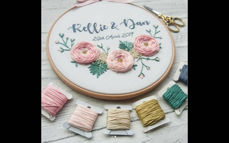 May include: Embroidered hoop art with pink flowers, green leaves, and the text "Kellie & Dan 26th April 2019".  The hoop is surrounded by spools of embroidery floss in pink, beige, gold, green, and blue.