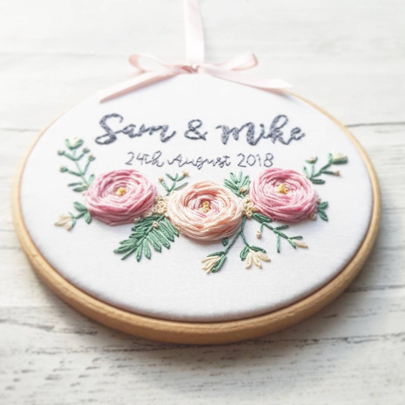 May include: Embroidered hoop with the names "Sam & Mike" and the date "24th August 2018" in cursive script. The hoop is decorated with three pink flowers and green leaves.