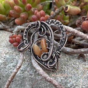 Könnte beinhalten: Ein handgefertigter Kupferdraht-Anhänger mit herzförmigem Design, mit einem zentralen orange-braunen Stein. Der Anhänger ist aufwendig mit dunklem Kupferdraht gewebt und zeigt einen einzigartigen, künstlerischen Stil. Der Anhänger wird auf einem natürlichen Hintergrund aus Sukkulenten präsentiert.