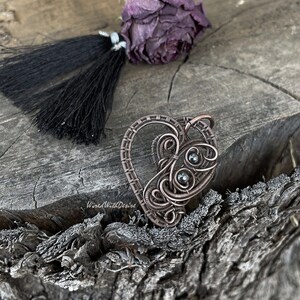 May include: A handcrafted copper wire heart pendant with intricate swirls and dark gray beads. The pendant is on a textured wooden surface, with a dried purple rose and black tassel. The text "WiredWithDesire" is visible.