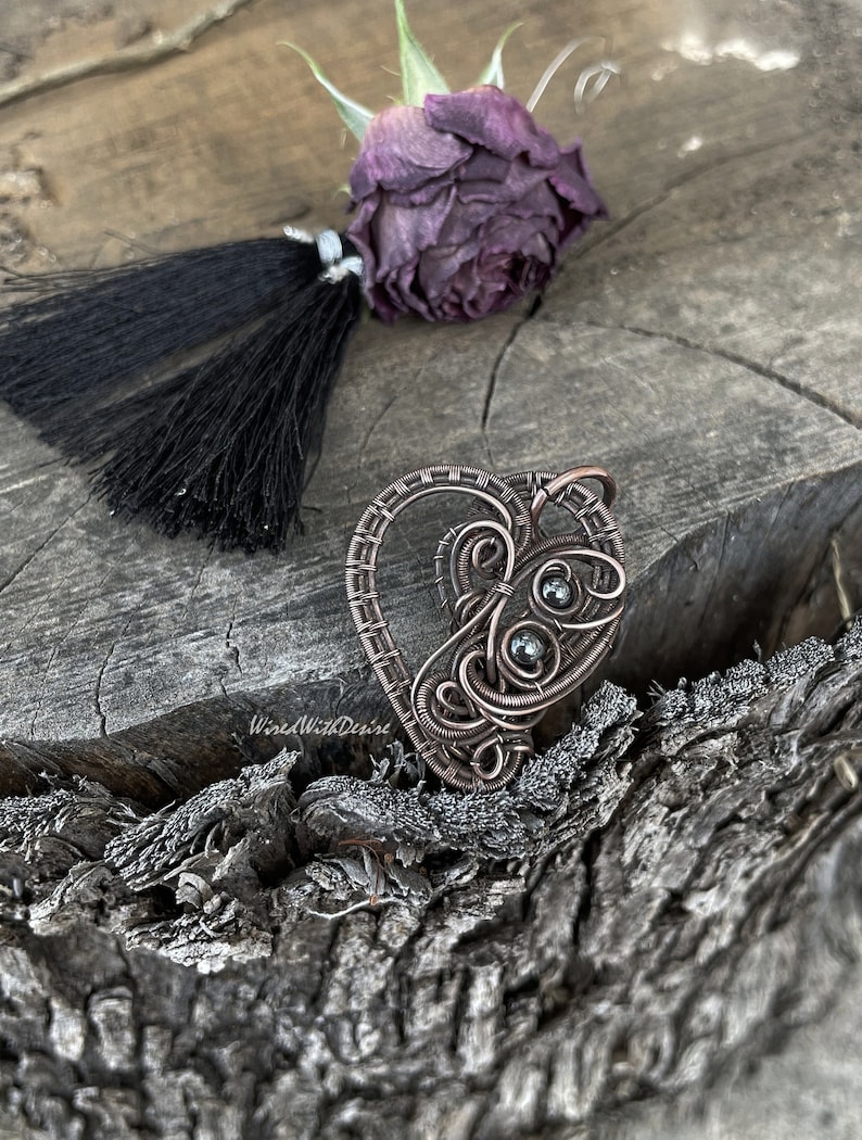 May include: A handcrafted copper wire heart pendant with intricate swirls and dark gray beads. The pendant is on a textured wooden surface, with a dried purple rose and black tassel. The text "WiredWithDesire" is visible.