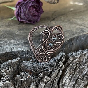 May include: A handcrafted copper wire pendant in a heart shape, featuring intricate wire wrapping and small, dark green beads. The pendant has a rustic, handmade aesthetic. The background includes a dried purple rose and a weathered wooden surface.