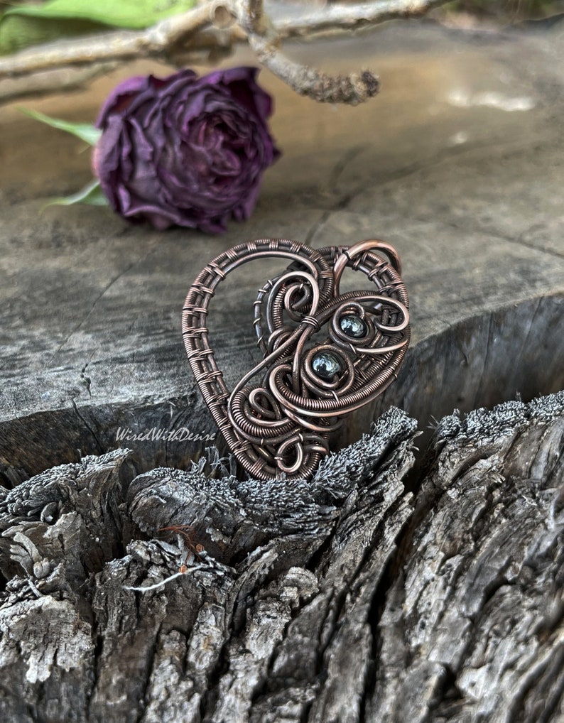 May include: A handcrafted copper wire pendant in a heart shape, featuring intricate wire wrapping and small, dark green beads. The pendant has a rustic, handmade aesthetic. The background includes a dried purple rose and a weathered wooden surface.