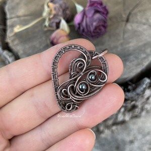May include: A copper-colored wire-wrapped pendant in a heart shape, featuring intricate swirls and three small, dark beads. The pendant is held in a hand, with the text "WiredWithDesire" visible. The background includes dried purple flowers.