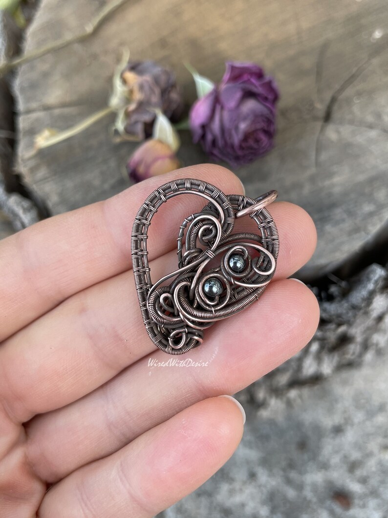May include: A copper-colored wire-wrapped pendant in a heart shape, featuring intricate swirls and three small, dark beads. The pendant is held in a hand, with the text "WiredWithDesire" visible. The background includes dried purple flowers.