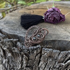 May include: A copper-colored wire-wrapped heart pendant with two dark gray beads. The pendant is on a weathered wooden surface, with a black tassel and a dried purple rose in the background. The pendant is handmade.