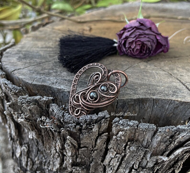 May include: A copper-colored wire-wrapped heart pendant with two dark gray beads. The pendant is on a weathered wooden surface, with a black tassel and a dried purple rose in the background. The pendant is handmade.