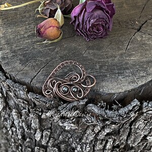 May include: A handcrafted copper wire heart pendant with intricate swirls and two small, dark green stones. The pendant rests on a weathered wooden surface, with dried roses in the background. The pendant is a unique piece of jewelry.