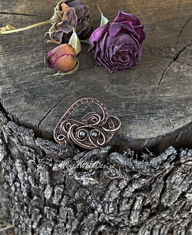 May include: A handcrafted copper wire heart pendant with intricate swirls and two small, dark green stones. The pendant rests on a weathered wooden surface, with dried roses in the background. The pendant is a unique piece of jewelry.