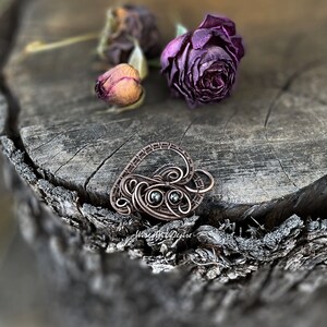 May include: A handcrafted copper wire pendant in a heart shape, featuring two small, dark gemstones. The pendant rests on a weathered wooden surface, with dried roses in the background. The pendant is a unique piece of jewelry.