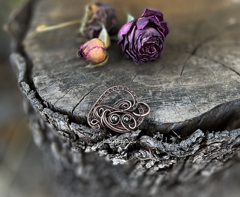 May include: A handcrafted copper wire pendant in a heart shape, featuring two small, dark gemstones. The pendant rests on a weathered wooden surface, with dried roses in the background. The pendant is a unique piece of jewelry.