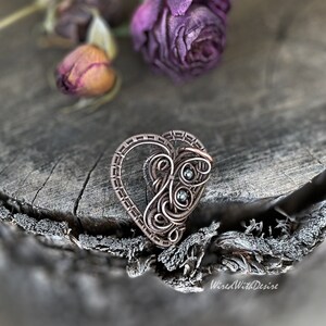 May include: A copper-toned wire-wrapped heart pendant with intricate detailing and two small, round, green gemstones. The pendant is set against a rustic wooden surface with dried purple roses, evoking a vintage or handcrafted aesthetic.
