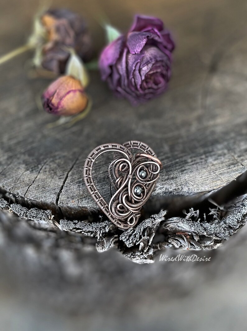 May include: A copper-toned wire-wrapped heart pendant with intricate detailing and two small, round, green gemstones. The pendant is set against a rustic wooden surface with dried purple roses, evoking a vintage or handcrafted aesthetic.