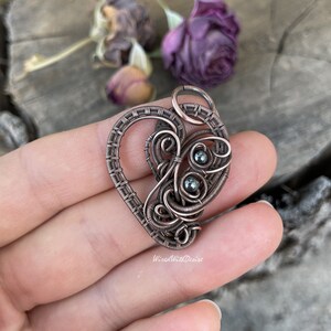 May include: A handcrafted copper wire pendant in a heart shape, featuring intricate wire wrapping and small, dark gray beads. The pendant is held in a hand, with the text "WiredWithDesire" visible. The background includes dried purple flowers.