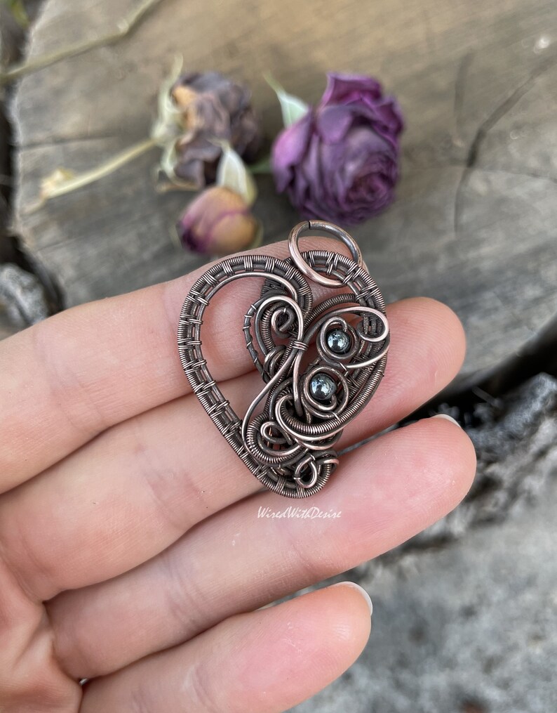 May include: A handcrafted copper wire pendant in a heart shape, featuring intricate wire wrapping and small, dark gray beads. The pendant is held in a hand, with the text "WiredWithDesire" visible. The background includes dried purple flowers.