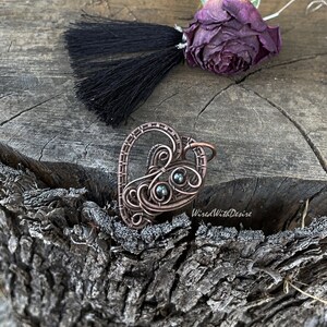 May include: A handcrafted copper wire pendant in the shape of a heart, featuring intricate wirework and dark, round beads. A black tassel and a dried purple rose are also present. The pendant is displayed on a textured wooden surface.