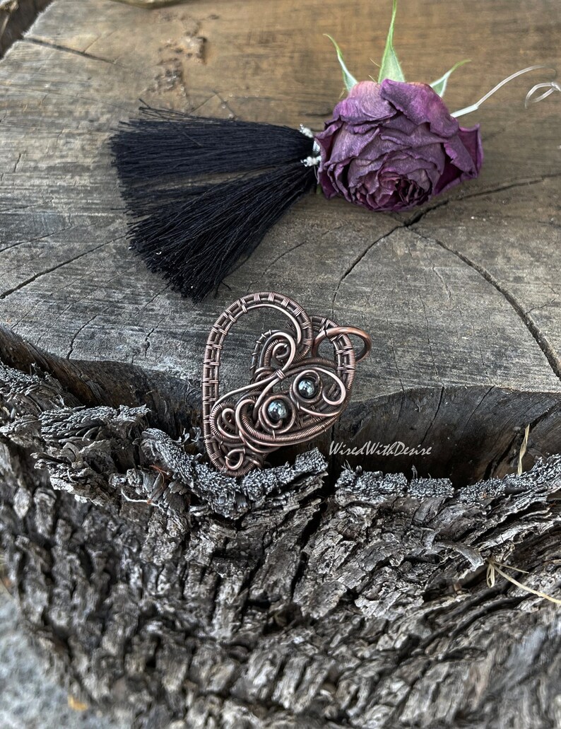 May include: A handcrafted copper wire pendant in the shape of a heart, featuring intricate wirework and dark, round beads. A black tassel and a dried purple rose are also present. The pendant is displayed on a textured wooden surface.