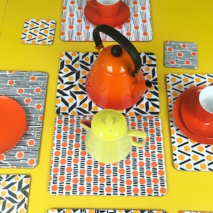 May include: A set of six orange, black and white patterned coasters with a red and yellow teapot and a red teacup on top of them. The coasters are on a yellow surface.