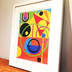 May include: Abstract geometric art print in a white frame. The print features a colorful design with circles, squares, and triangles in shades of red, yellow, blue, green, and black.
