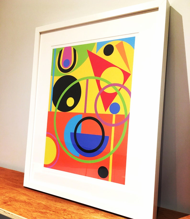 May include: Abstract geometric art print in a white frame. The print features a colorful design with circles, squares, and triangles in shades of red, yellow, blue, green, and black.