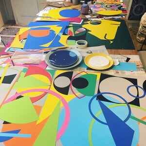 May include: A table covered in colorful paper shapes, including circles, squares, and triangles. The shapes are in various colors, including yellow, blue, green, red, and pink. The table is covered in newspapers and magazines, and there are some tools and supplies scattered around.
