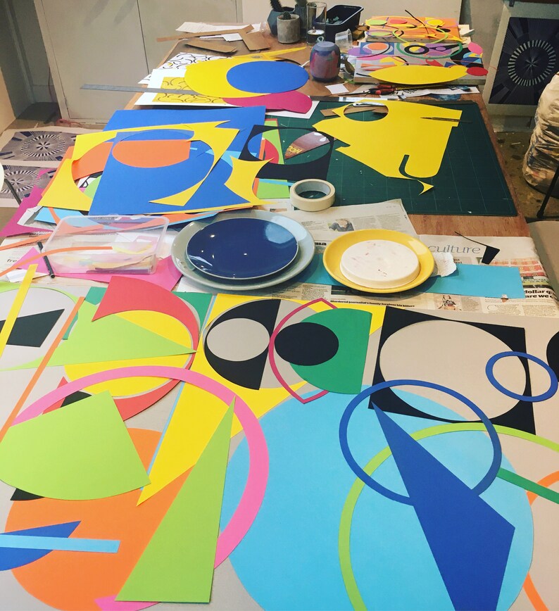 May include: A table covered in colorful paper shapes, including circles, squares, and triangles. The shapes are in various colors, including yellow, blue, green, red, and pink. The table is covered in newspapers and magazines, and there are some tools and supplies scattered around.
