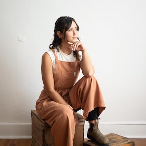 May include: A terracotta-coloured jumpsuit worn over a white t-shirt. The jumpsuit features a square pocket and a small logo patch. The person is wearing brown boots and sitting on a wooden crate. The background is a plain white wall.