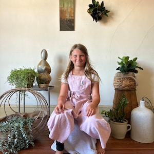 May include: A young person wearing a pink linen overall dress sits on a white cushion. The person is smiling and looking at the camera. There are plants and a ceramic sculpture in the background.