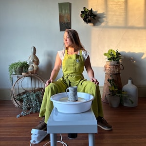 Könnte beinhalten: Eine Frau in einer grünen Schürze sitzt an einer Töpferscheibe mit einer Tasse auf der Scheibe. Die Scheibe steht auf einem weißen Tisch mit einem Fußpedal. Im Hintergrund befinden sich Pflanzen und eine Skulptur.