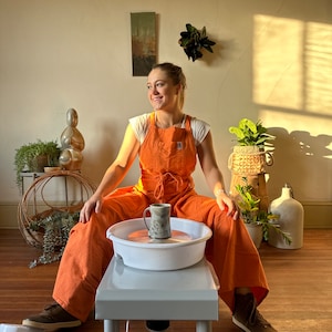 Könnte beinhalten: Eine Frau in einem orangefarbenen Overall sitzt vor einer Töpferscheibe und hält eine Keramiktasse. Die Scheibe ist weiß und hat einen grauen Sockel. Die Frau lächelt und schaut nach rechts.