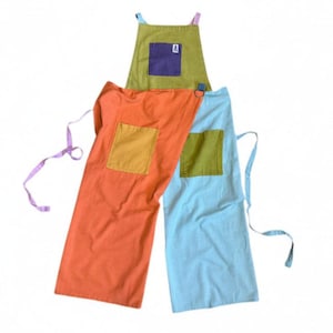 Pode incluir: Um avental de blocos de cores com um peitilho verde-azeitona com um bolso roxo e a letra A, uma secção laranja com um bolso amarelo e uma secção azul-clara com um bolso verde-azeitona. O avental inclui fitas de tecido finas em ambos os lados.