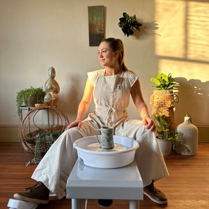 May include: A woman wearing a light brown jumpsuit and a white apron sits at a pottery wheel. She is holding a ceramic mug in her hand. The pottery wheel is white and has a gray base. The woman is looking to the right of the image.