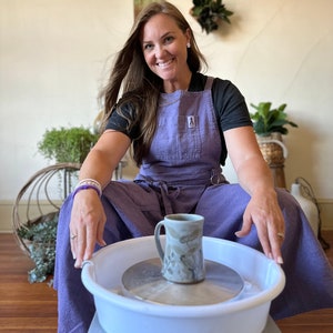 May include: A woman wearing a purple apron sits in front of a pottery wheel with a white basin. A ceramic mug with a brown and white design sits on the wheel's platform.