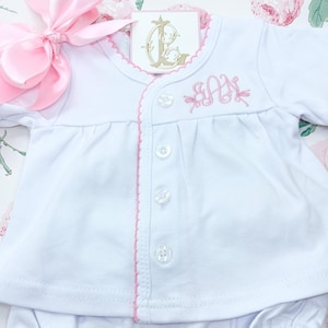 May include: White baby outfit set with pink trim and a matching pink bow. The set includes a button-down top with embroidered initials and ruffled bloomers. The outfit is displayed on a floral background.
