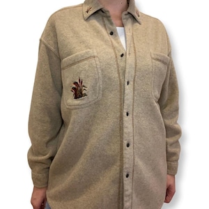 Peut inclure: Une chemise beige à manches longues avec un devant boutonné et un motif brodé brun sur la poche gauche. La chemise a un col et une bordure brune autour du col et des poches.
