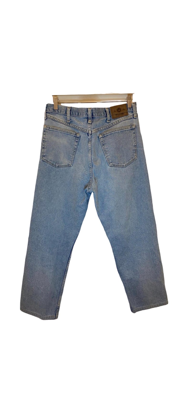 Peut inclure: Un jean en denim bleu clair avec un &eacute;cusson en cuir brun sur la poche arri&egrave;re qui dit "Wrangler".