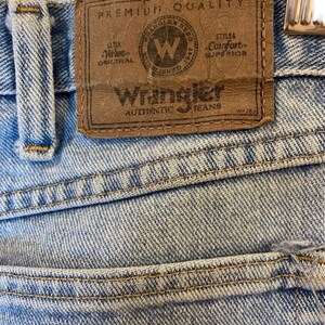 Peut inclure: Un gros plan sur un jean en denim bleu clair avec un patch en cuir brun qui indique "Premium Quality" et "Wrangler Authentic Jeans".