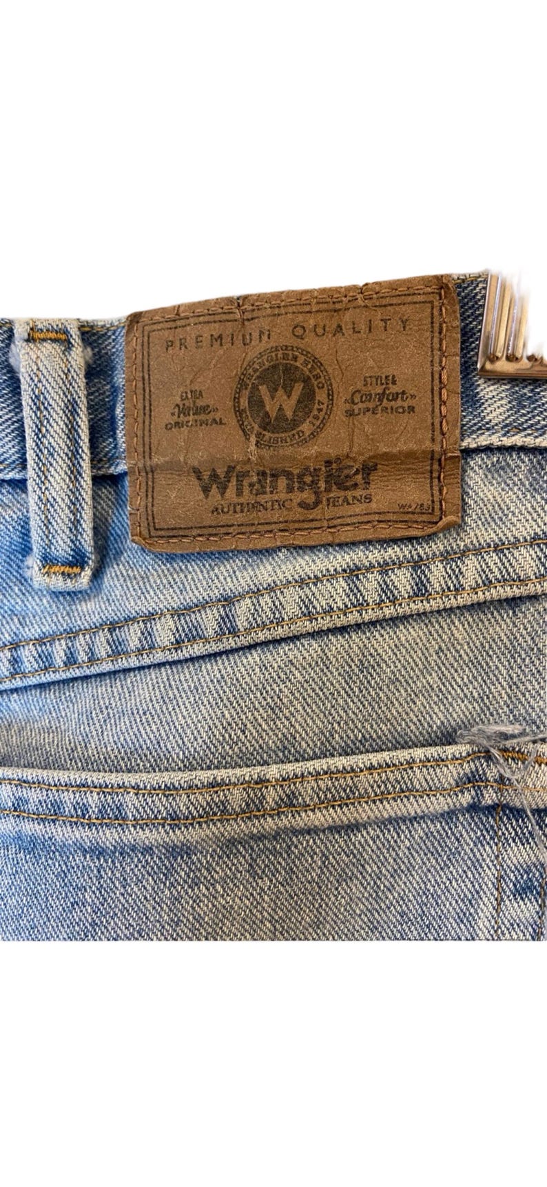 Peut inclure: Un gros plan sur un jean en denim bleu clair avec un patch en cuir brun qui indique "Premium Quality" et "Wrangler Authentic Jeans".