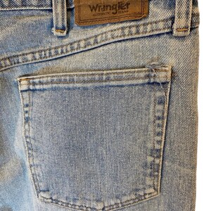 Peut inclure: Gros plan sur la poche arri&egrave;re d'un jean bleu clair. Le jean a un patch en cuir marron avec le texte "Premium Quality" et "Wrangler Authentic Jeans".