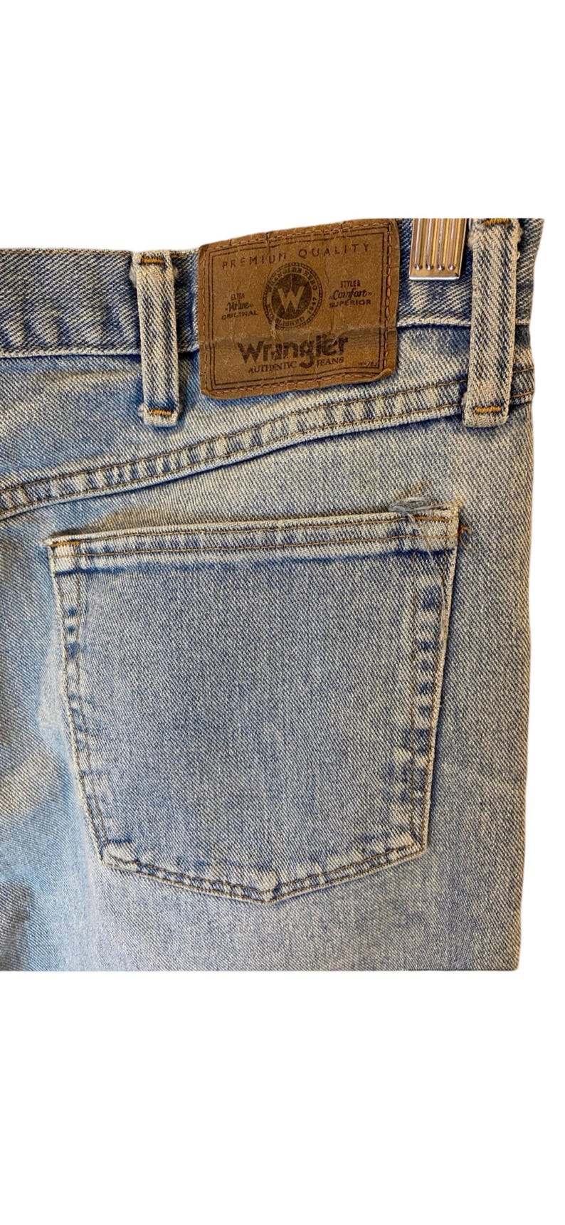 Peut inclure: Gros plan sur la poche arri&egrave;re d'un jean bleu clair. Le jean a un patch en cuir marron avec le texte "Premium Quality" et "Wrangler Authentic Jeans".
