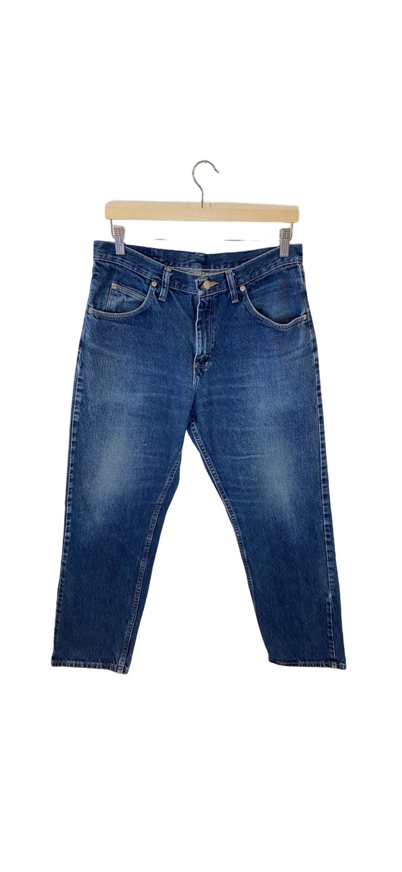 May include: A pair of blue denim jeans hanging on a wooden hanger. The jeans have a classic straight leg style and a button fly.