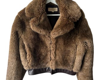 Puede incluir: Chaqueta de piel sintética marrón con cuello ancho y ribete de cuero marrón oscuro en la cintura y los puños. La chaqueta está colgada de una percha blanca sobre un fondo blanco. La chaqueta tiene una etiqueta en el interior.