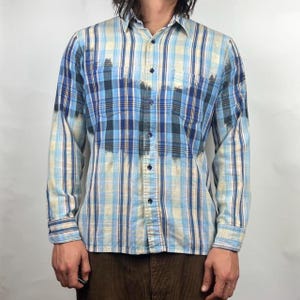 May include: A long-sleeved button-down shirt with a blue, white, and tan plaid pattern. The shirt has a faded, distressed appearance with darker blue patches. It features two chest pockets and a classic collar. The shirt is made of a lightweight fabric.